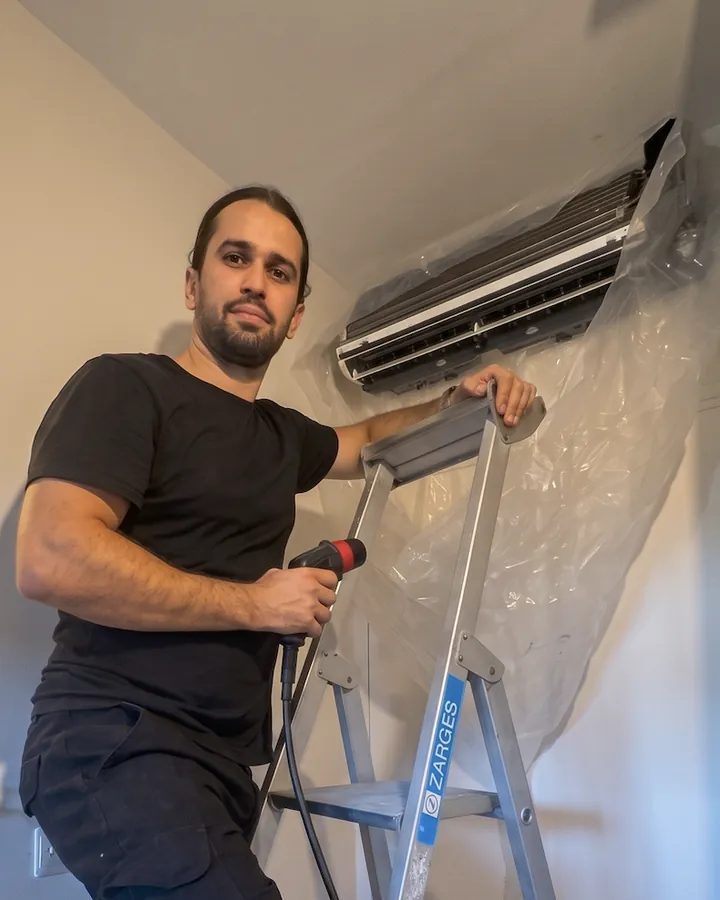 Dorian Olsak, responsable technique de Seven Clim, sur son escabeau devant une unité intérieure bâchée, lance de rinçage en main
